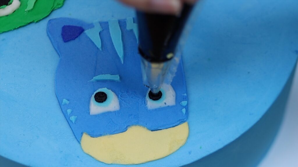 piping the eyes and facial details onto catboy pj masks for a character cake with no fondant