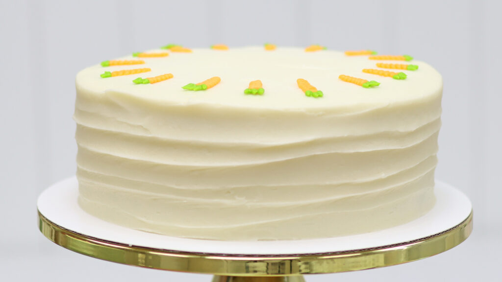A finished carrot cake with cream cheese frosting sitting on a cake stand, ready to serve. The cake has a textured frosting finish and decorative buttercream carrots on top.