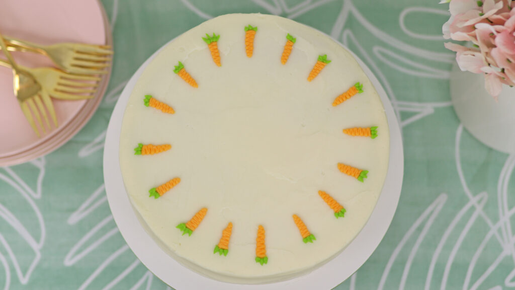 A finished carrot cake with cream cheese frosting sitting on a cake stand, ready to serve. The cake has a textured frosting finish and decorative buttercream carrots on top.